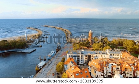 Drone photo captures Kołobrzeg's maritime charm, featuring the iconic lighthouse, cerulean sea, turbulent waves, a distant pier, and autumnal hues on the trees.