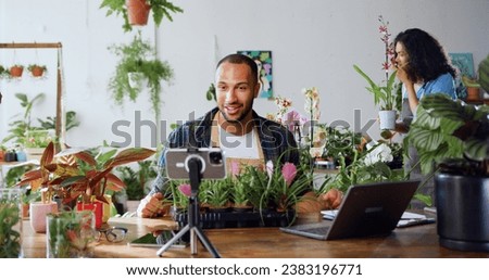 Handsome man gardener in apron using phone and recording blog video with clients. Young adult content creator influencer blogger recording a video talking about indoor plants care from workplace.