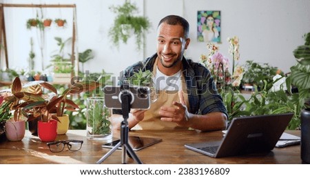 Gardener african american man blogger using phone while caring indoor plants and use a shovel on table. Concept of plants care and small bussines garden. Spring planting. Social media.