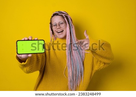 Excited young woman with braids pointing finger to blank phone green screen with copy space for app, isolated on yellow background. High quality photo