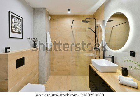 Decorated modern bathroom in boho style with a walk-in cabin, rain shower column and black built-in cabinets, with honey-colored wood-imitating tiles and round led light mirror Royalty-Free Stock Photo #2383253083