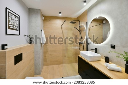 Decorated modern bathroom in boho style with a walk-in cabin, rain shower column and black built-in cabinets, with honey-colored wood-imitating tiles and round led light mirror Royalty-Free Stock Photo #2383253085