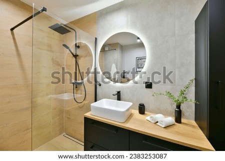 Decorated modern bathroom in boho style with a walk-in cabin, rain shower column and black built-in cabinets, with honey-colored wood-imitating tiles and round led light mirror Royalty-Free Stock Photo #2383253087