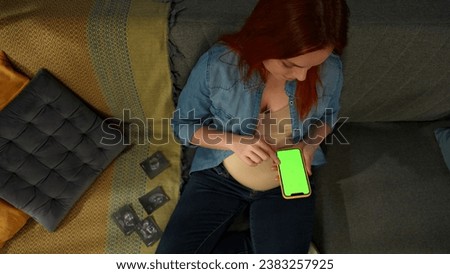 A close up shot shows a pregnant woman sitting on a sofa. She is holding a phone with a green screen in her hands. Nearby are ultrasound imaging. She smiles and is happy. Place for advertising