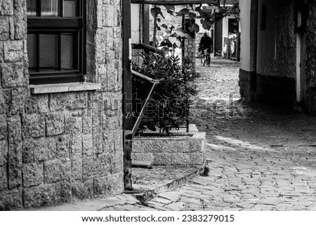 Old man riding a bicycle, in the 
alley street of Ohrid City. Sunrise. Morning sun.  Wooden window frame, part of the building architecture. Black and white colors. Monochrome. Macedonia 2023. Royalty-Free Stock Photo #2383279015