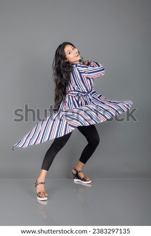 A beautiful, young Indian woman wearing black leggings, black top and red, blue, white striped tunic. She is laughing, twirling,  jumping and dancing in joy for celebration in a studio