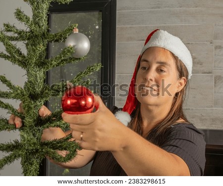 a woman in the foreground starting to assemble the Christmas tree, placing red and silver decorative balls. The woman has a black dress and a Santa Claus hat.