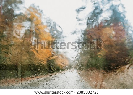 Autumn blurry background picture through wet window. No people image with yellow, green and orange trees. View from car with raindrop texture. Scenic road