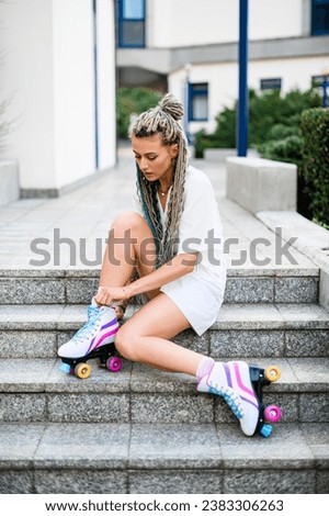 Beautiful girl with braids in hair sitting on stairs and tying laces on retro fashioned colorful roller skates, front wiew. Blurred street background. Concept: sports, extreme, healthy lifestyle.