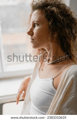 Portrait of curly woman in top and crochet cover-up standing near window in studio 