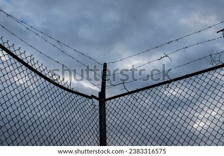 Close up photo of a barbed wire chain link fence against a dark stormy sky Royalty-Free Stock Photo #2383316575