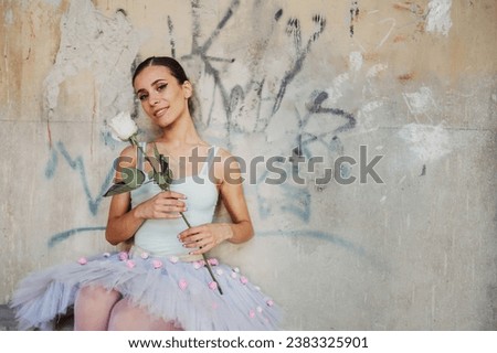 Portrait of an elegant female ballerina dancer wearing ballet skirt with pink flowers posing with white rose in her hands while showing incredible physical abilities. Looking into the camera.