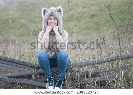 A girl in a wolf hat sits on a wooden fence in the village. Autumn