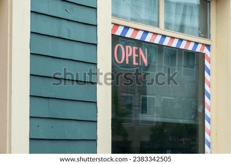 The exterior of a green wooden building with cream colored trim. The shop has an illuminated red open sign hanging in the glass window of the barbershop. There's a red, white and blue striped edging. 