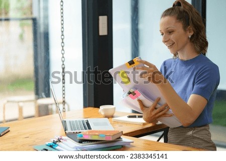 A female freelance holds documents to read business data and financial charts while working with a laptop at an office table.