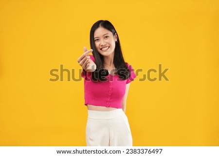 Photo of beautiful young asian girl in yellow sweater, smiling and showing finger hearts, standing happy against yellow background.
