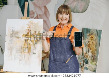 Elderly artist woman 50 years old wears casual clothes stand near easel with painting artwork point on blank screen mobile cell phone spend free spare time in living room indoor. Leisure hobby concept