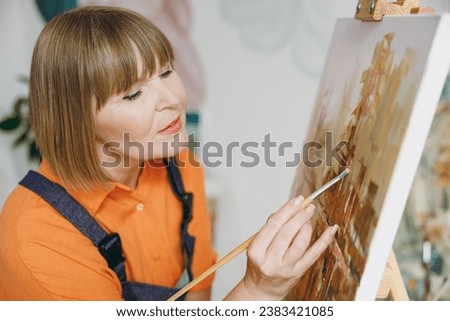 Close up elderly artist woman 50 years old wears casual clothes sit near easel with painting artwork paint do acrylic brush stroke spend free spare time in living room indoor. Leisure hobby concept