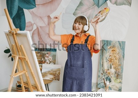 Elderly happy artist woman 50 years old wears casual clothes stand near easel with painting artwork paint listen music in headphones spend free spare time in living room indoor. Leisure hobby concept