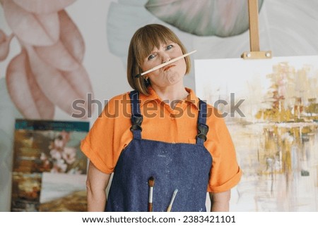 Elderly artist woman 50 years old wearing casual clothes stand near easel with painting hold brush on lips pov mustache look camera spend free spare time in living room indoor. Leisure hobby concept