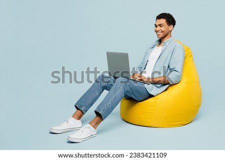 Full body young IT man of African American ethnicity wear shirt casual clothes sit in bag chair hold use work on laptop pc computer isolated on plain pastel light blue cyan background studio portrait