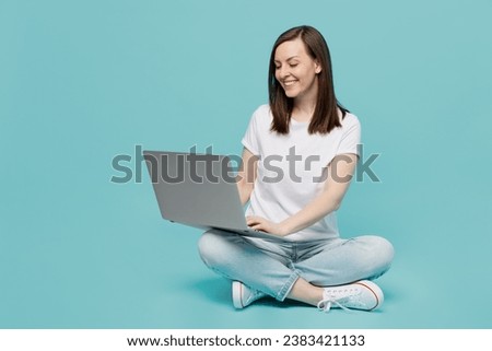 Full body young fun caucasian woman 20s she wear white t-shirt sitting on floor hold use work on laptop pc computer isolated on plain pastel light blue cyan background studio. People lifestyle concept