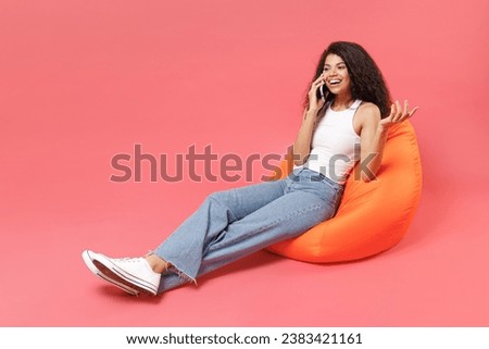 Full body young fun smiling happy woman of African American ethnicity wear casual white tank shirt sitting in orange bean bag chair talk by mobile cell phone spread hand isolated on pink background Royalty-Free Stock Photo #2383421161