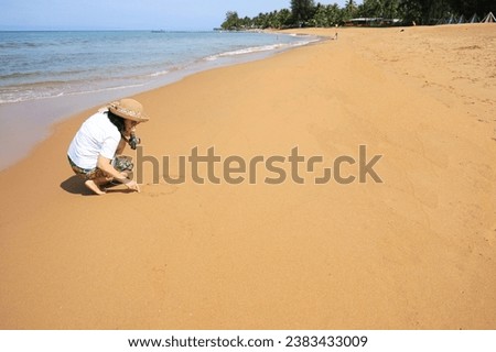 A woman sits with her back to the sea and is writing the letters "L" "O" to express the word LOVE on the fine sand. Female tourists drawing in the sand happily at the beach while visiting the sea. Royalty-Free Stock Photo #2383433009