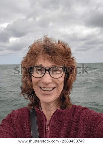 Mature woman takes a smiling selfie for social media at Mordialloc pier overlooking the sea. She is windswept and not wearing any make up.