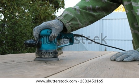 carpenter's hands holding a sander and polishing the surface of brownish solid wood outdoors, processing wood texture with a sanding tool, smoothing out unevenness of wood material with sandpaper