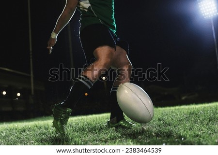 Rugby player drops the ball on the ground and then kicks it just as it bounces. Rugby player hitting a dropped goal under lights at sports arena. Royalty-Free Stock Photo #2383464539