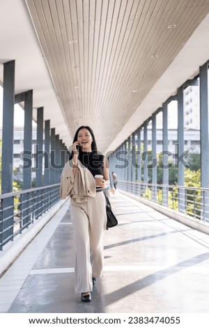 A busy and confident millennial Asian businesswoman is talking on the phone with someone while walking on a skywalk. Urban busy life concept Royalty-Free Stock Photo #2383474055