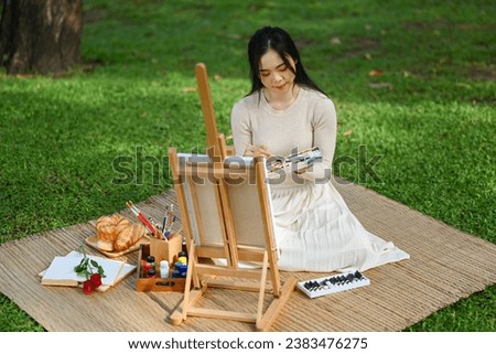 Peaceful young Asian woman artist holding palette painting a picture on an easel at the park.