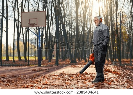 Gray haired smiling male in gloves standing, cleaning vacuuming fallen leaves in park. Side view of caucasian handyman working outdoors in autumn, against basketball backboard. Seasonal work concept.