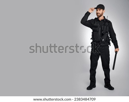 Smiling male police officer in uniform standing with baton in studio. Front view of bearded policeman holding truncheon and looking at copy space beside, on gray background. Concept of work, tool.