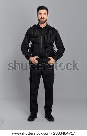 Happy police officer with dark hair posing, with hands on belt in studio. Front view of attractive cop in full gear smiling to camera, on gray background. Concept of work, profession, police.