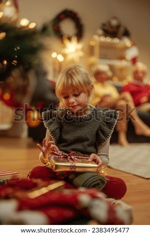 Cute little girl is sitting on the floor and packing and decorating Christmas present in front of a Christmas tree and smiling at the camera, while her grandparents are in the background