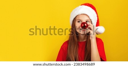 Happy little girl holding red christmas tree ball in front of his nose, wearing Santa hat posing against yellow studio wall background