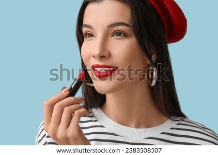 Beautiful young woman in beret applying red lipstick on blue background