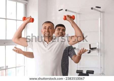 Mature man training with dumbbells and therapist in rehabilitation center