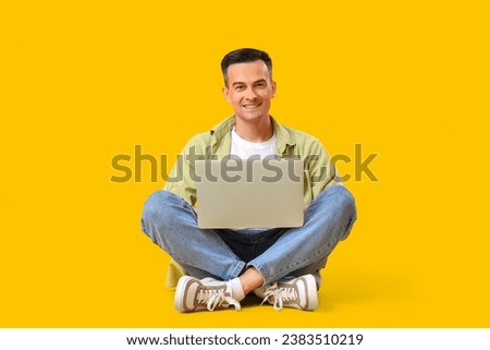 Happy young man sitting on floor with laptop against yellow background