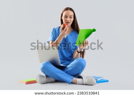 Female medical intern with apple studying on light background