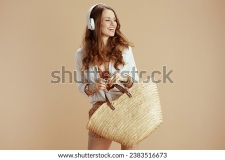 Beach vacation. smiling elegant middle aged housewife in white blouse and shorts isolated on beige with straw bag and headphones.