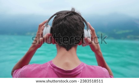 Back of young boy putting headphones over ear while standing in lake shire view. Teenager listening to music, podcast, or audiobook, contemplating nature