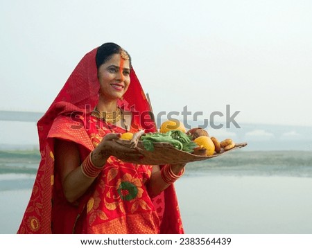 A woman offering prasad, fruits, vegetables, and other items and a Diya to pray sun God at a lake during Chhath Puja. Hindu devotees worship God sun with a ritual while standing in a river - Hindu ... Royalty-Free Stock Photo #2383564439