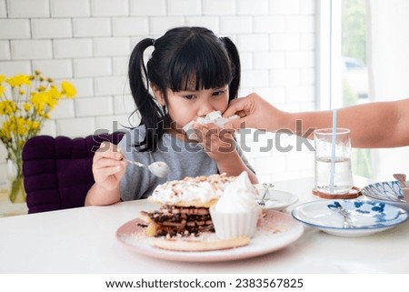 Mother's hand is using a tissue to clean her asian daughter's mouth from food stains while eating at the table. sweet cake in cafe shop