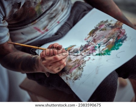 From above of crop unrecognizable person with arms dirty of colorful blots wearing paint stained t shirt holding paintbrush and drawing multicolor picture