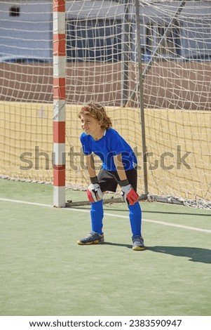 Full body of preteen goalkeeper in activewear leaning on knees and looking forward while playing soccer during training on sports ground