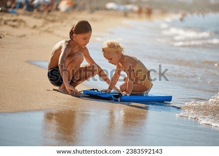 Side view of boy and girl in swimsuits sitting on sandy seashore and playing with blue surfboard together during summer vacation