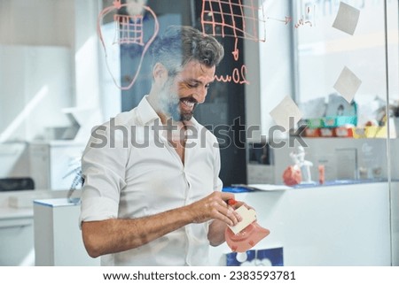 Through window of glad male engineer in formal wear with respirator taking sticky notes while standing near glass wall with written information